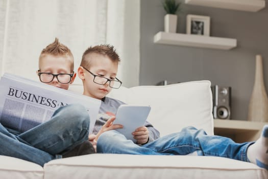 Two young boys wearing spectacles relaxing on a sofa reading the business news and an e-book on a tablet with engrossed expressions