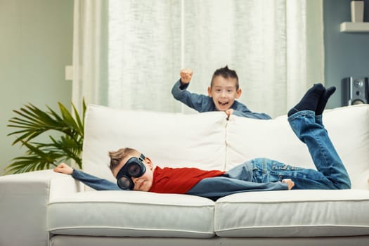 Two mischievous young brothers having fun playing on a sofa with one shouting from the rear as the second lies on his stomach wearing goggles