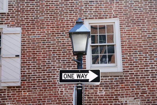 One way sign hanging on a black pole with street lamp, with a traditional brick wall on the back with white window