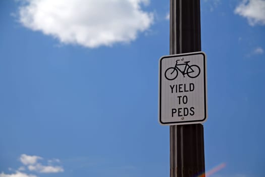 Yield to Peds sign hanging on a black pole with the sky and the clouds on the back, with bicycle icon