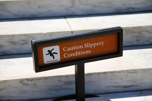 Caution, slippery conditions brown sign with icon representation on marble steps with a black holder on a sunny day
