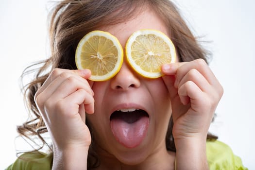 Mischievous little girl holding sliced lemons to her eyes while sticking out her tongue at the camera isolated on white