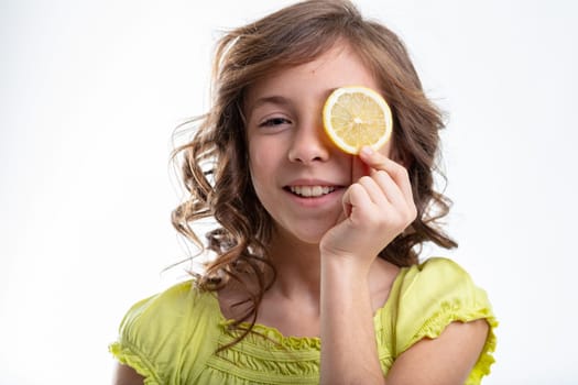 Cute little girl with curly hair holding a slice of lemon to one eye with a happy friendly smile in a head and shoulders portrait on white