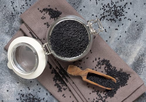 Indian spice Black cumin (nigella sativa or kalonji) seeds glass jar with a wooden scoop, top view. Traditional asian medicine, healthy and vegetarian food concept