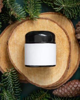Glass jar between fir branches and pine cones on wooden piece top view. Brand packaging mockup. Dark cream container for skincare product, cream or serum. Healthcare concept. Natural cosmetics.