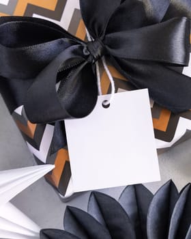 Present with blank square gift tag and black bow close up. Gift boxes wrapped in chevron geometric paper near black and white decor. Christmas, New Year, Birthday, Anniversary label mockup