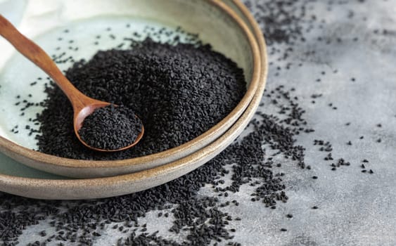 Plate of Indian spice Black cumin (nigella sativa or kalonji) seeds on grey table with a wooden spoon, close up with copy space. Traditional asian medicine, healthy and vegetarian food concept