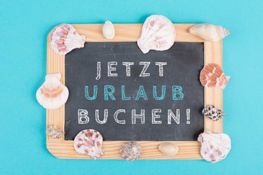 Book vacation now is standing in german language on the chalkboard, seashells bulit frame, holiday and summertime topic, travel concept