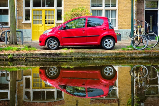 Red car on canal embankment in street of Delft with reflection and bicycle. Delft, Netherlands