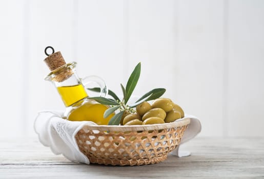 Olive oil bottle and olives with branch leaves in basket on wooden table