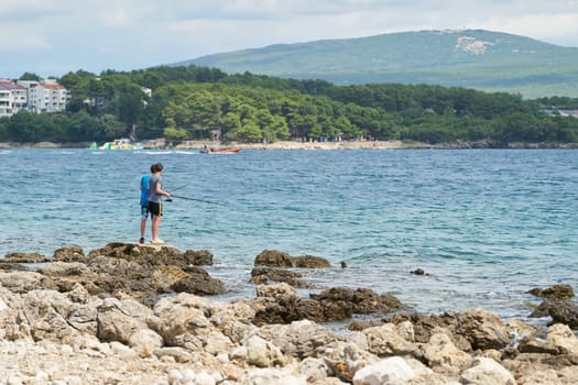 two children standing on rocky coast near town Krk in Croatia fishing