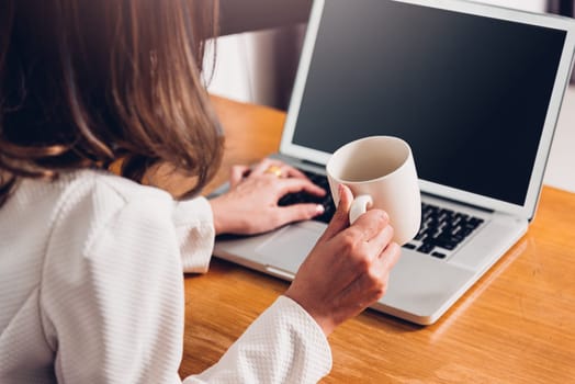 Beautiful business woman lifestyle working using laptop computer and holding coffee cup on hand while enjoying in modern desk office near window, work from home concept