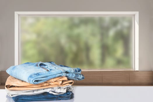 Stack of jeans. Closeup of a pile of colorful female denim pants on a bright table against abstract blurred natural windows background. Copy space.