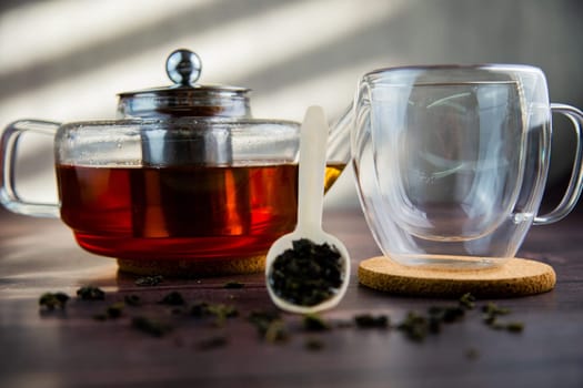 A wooden spoon with tea lies against a dark wood background, a mug and a teapot are visible from behind, a photo in dark colors. High quality photo