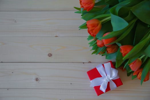 Pink tulips with green leaves lie in the upper corner on a wooden background, and next to it is a gift in a red package with a bow. High quality photo