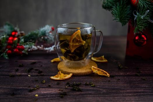 A transparent cup of tea stands on a dark table, on a wooden stand, dried orange slices float in the cup, citrus slices lie next to it, tea is scattered, a photo in a dark key. High quality photo