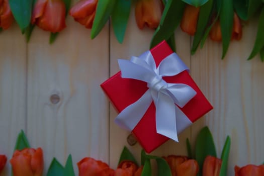 Pink tulips with green leaves lie in a row at the bottom and top on a wooden background, in the middle is a gift in a red package with a white bow. High quality photo