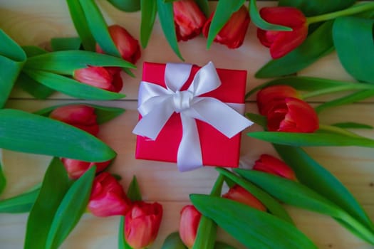 Pink tulips with green leaves lie in a circle on a wooden background, in the middle of Lesha's gift in a red package with a white bow. High quality photo
