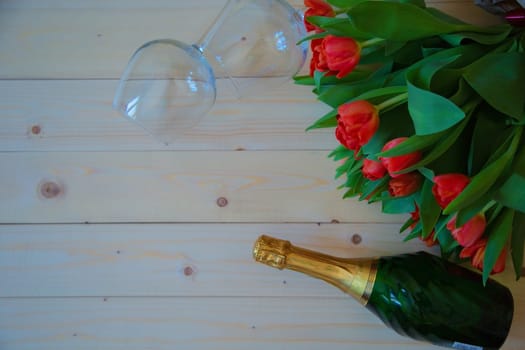 Pink tulips with green leaves lie in the upper corner on a wooden background, and on the sides of the bouquet are wine glasses and sparkling wine. High quality photo