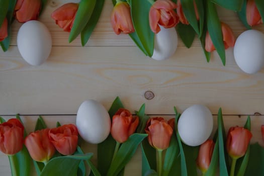 Pink tulips with green leaves lie in a row at the bottom and top on a wooden background with a place for text, white Easter eggs are cut next to them. High quality photo