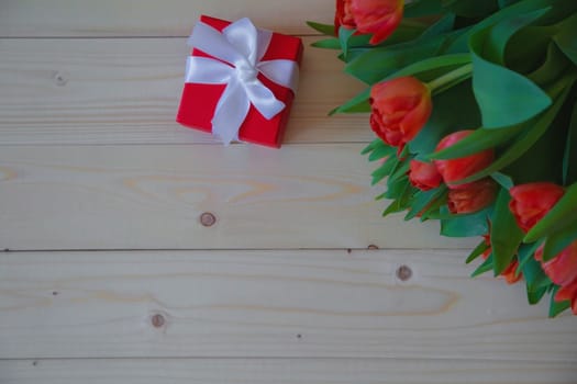 Pink tulips with green leaves lie in the upper corner on a wooden background, and next to it is a gift in a red package with a bow. High quality photo