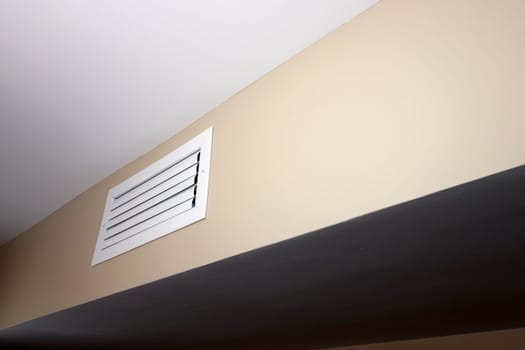 One closed air vent that is rectangle, white, small and situated in a beige wall near a white ceiling. A closed white rectangle air duct vent located in a home in a beige wall below a white ceiling.