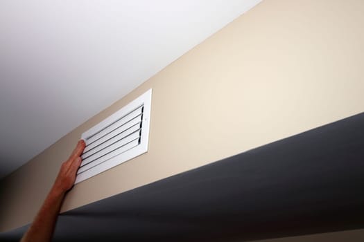 A left caucasian adult hand held up to a closed small air vent on a beige wall near a white ceiling. White metal rectangle furnace closed vent grid with a left hand on top of the left side close-up.