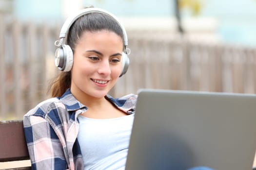 Happy teen wearing headphones using laptop on bench