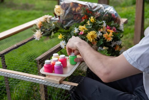 Adult arts and crafts art therapy creativity concept, man paints silk flowers in calm outdoor setting with green grass background and crutches leaning against a post. Shot in natural light with copy space.