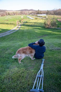 Crutches in the foreground of this idyllic rural nature background scene with a young man and his loving golden retriever dog laying on a grassy hill in natural golden hour sunlight one person and copy space.