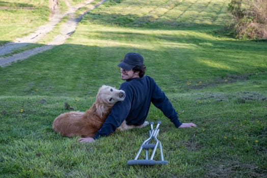 Dog cuddles with his owner on a rural grassy hill with a pair of crutches on the ground in the foreground. Dog looks back toward the camera in outdoor natural golden hour sunlight with nature background one person and copy space.
