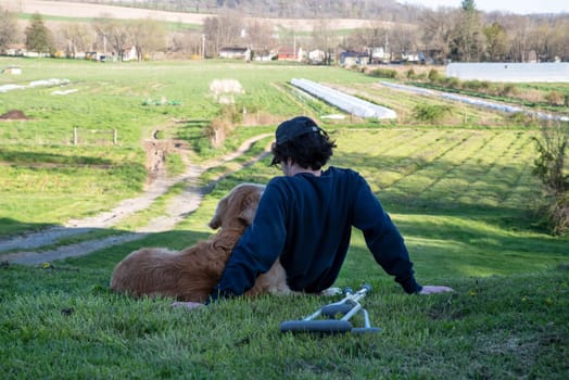 Crutches in the foreground of this idyllic rural farm background scene with a young man and his loving golden retriever dog cuddling with an arm around each other sitting on a grassy hill in natural golden hour sunlight one person and copy space.