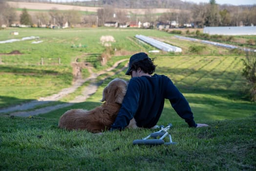 Crutches in the foreground of this idyllic rural farm background scene with a young man and his loving golden retriever dog cuddling and looking at each other sitting on a grassy hill in natural golden hour sunlight one person and copy space.