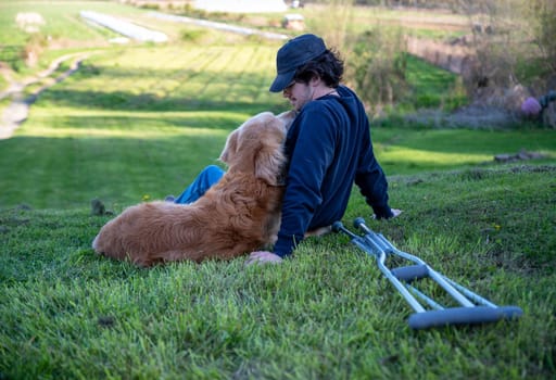 Crutches in the foreground of this idyllic rural farm background scene with a young man and his loving golden retriever dog cuddling with an arm around each other sitting on a grassy hill in natural golden hour sunlight one person and copy space.
