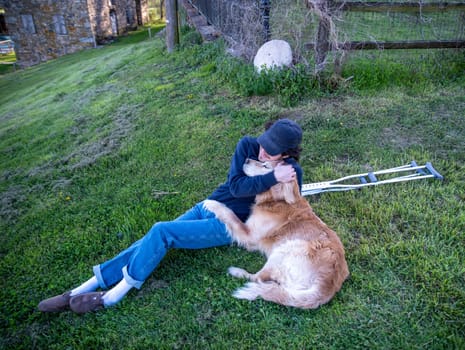 Crutches in the foreground of this idyllic rural farm background scene with a young man and his loving golden retriever dog cuddling with an arm around each other sitting on a grassy hill in natural golden hour sunlight one person and copy space.