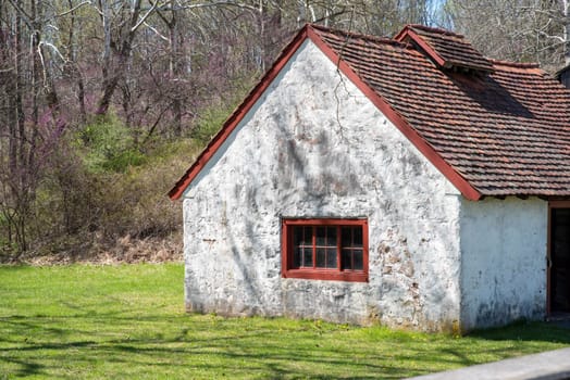 Charming idyllic colonial American Pennsylvania stone cottage with red window and peaked roof in golden hour sunlight. Green grass and woodland background with no people and copy space.