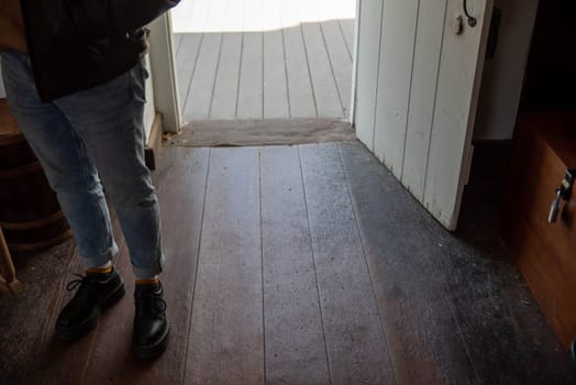 One person's feet stand inside the open door of an old colonial American home with old, hardwood floors and rustic antique door and stone threshold. Selective focus on foreground with copy space and wood grain texture.