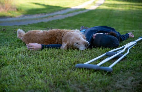 Crutches in the foreground of this idyllic rural nature background scene with a young man and his loving golden retriever dog laying on a grassy hill in natural golden hour sunlight one person and copy space.