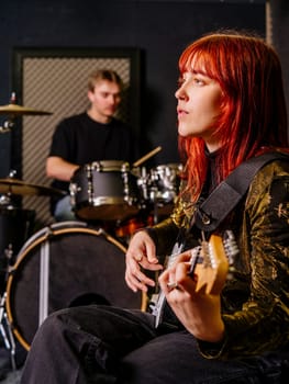 Young redhead woman playing electric guitar and a male drummer in a recording studio.