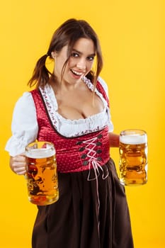 Beautiful waitress wearing traditional dirndl and holding two mass beer steins over yellow background.
