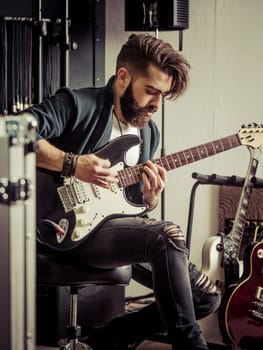 Young man with beard sitting and playing his electric guitar in a recording studio.