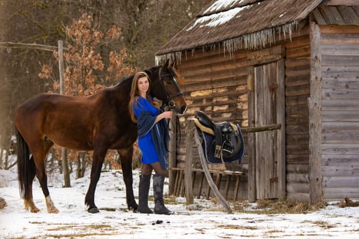 A beautiful girl in a short blue dress stands with a horse from an old wooden house