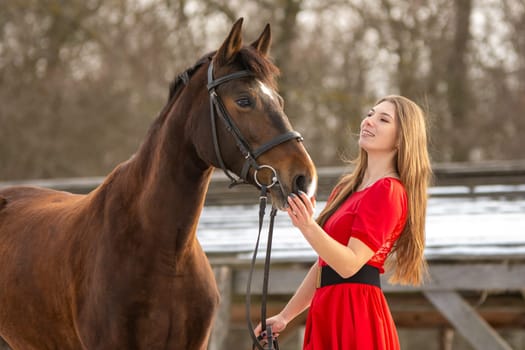 A beautiful girl in a red dress strokes a horse in the rays of the setting sun