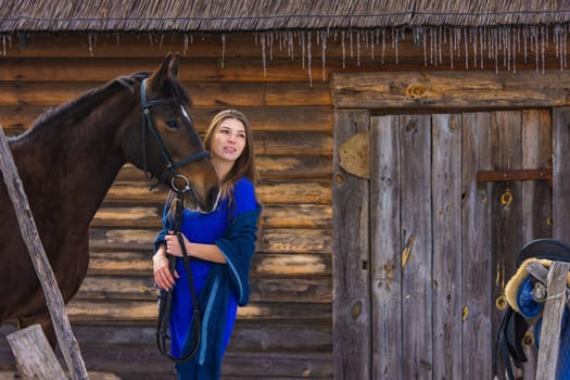 A beautiful young girl holds a horse by the bridle, against the background of a log wall