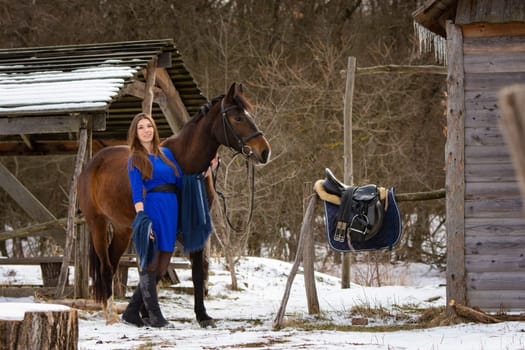 A beautiful girl in a short blue dress walks with a horse near the farm in winter