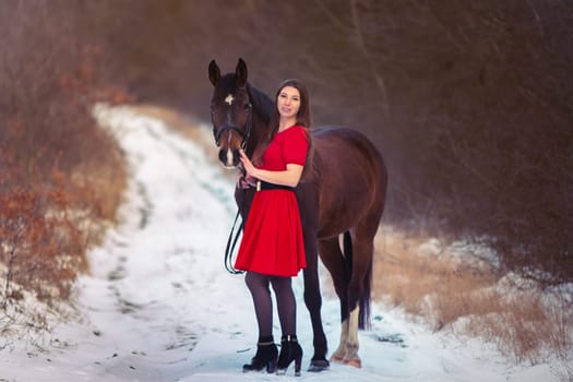 A beautiful girl in a red dress walks with a horse against the backdrop of a winter forest