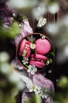 Top view on pink strawberry macaron cookies in bowl on wooden brown deck boards background, white apple blossom flowers. Beautiful colorful composition flat lay with sweet small French macaroon cakes