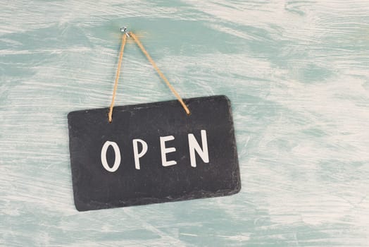 The word open is standing on a chalkboard sign, hanging on a blue textured weathered door, concept for business, stores and restaurants