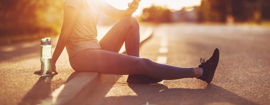Woman sitting on the border of the roadside with smartphone after a run. Panoramic view. Concept of the healthy lifestyle.