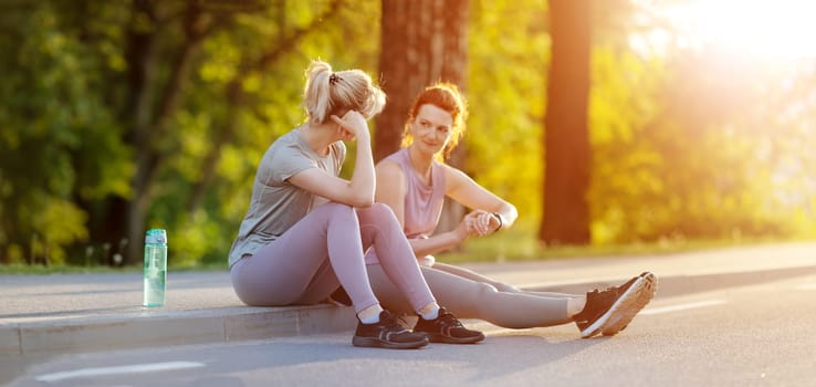 Two women sitting on the border of the roadside after a run. Panoramic view. Concept of the healthy lifestyle.